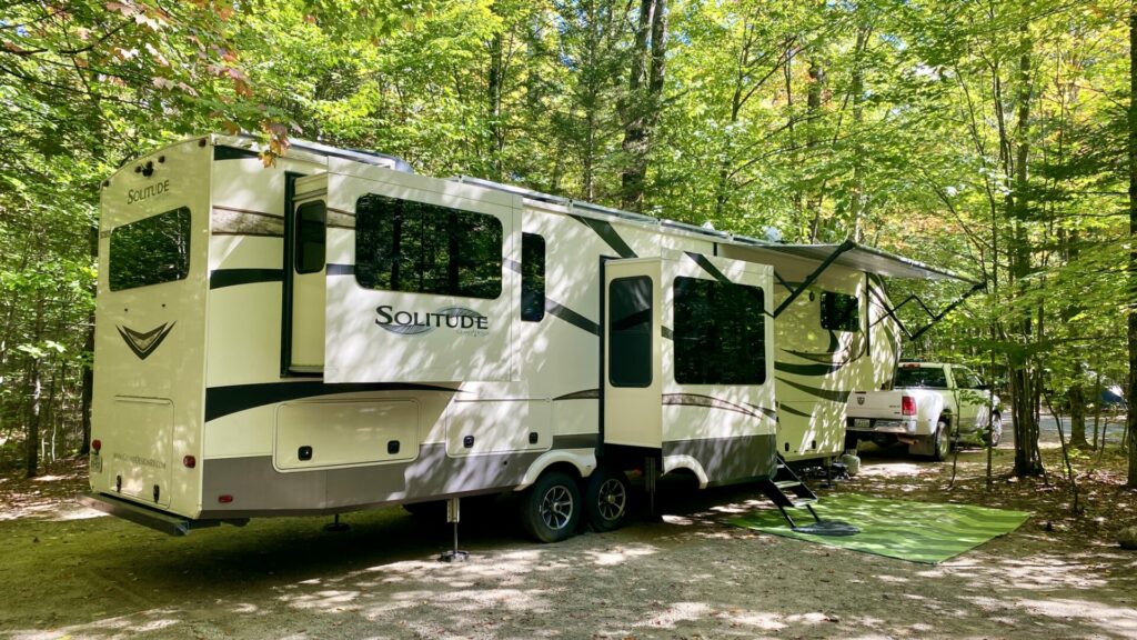 A fifth wheel RV parked in a campground with an outdoor rug on the ground and the awning out 