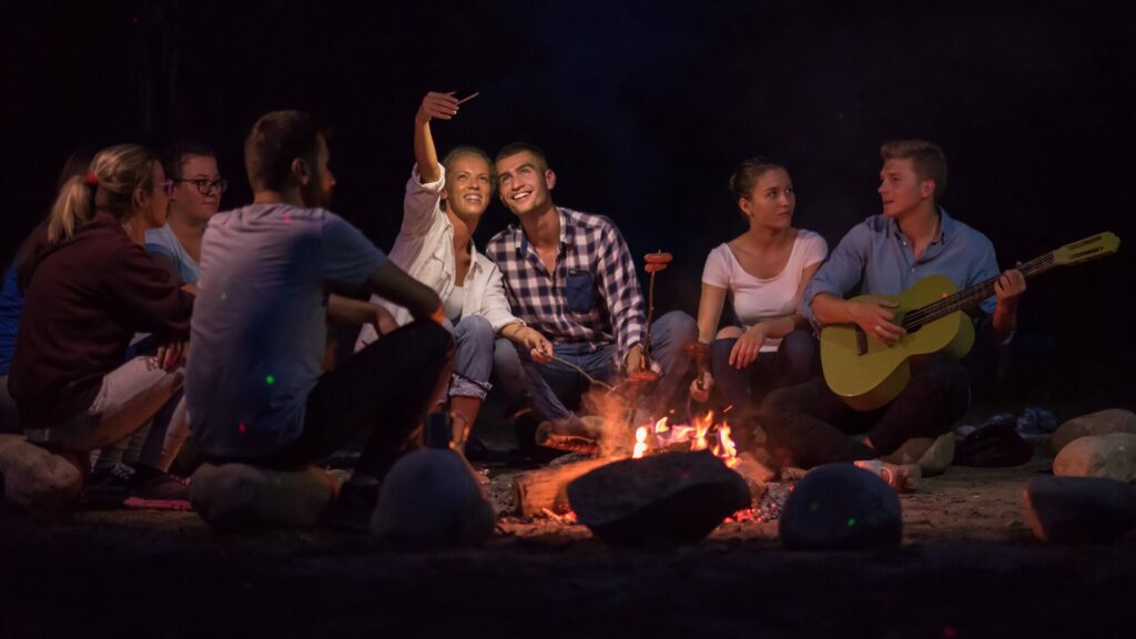 A group of people around a campfire taking pictures and playing on a guitar. 