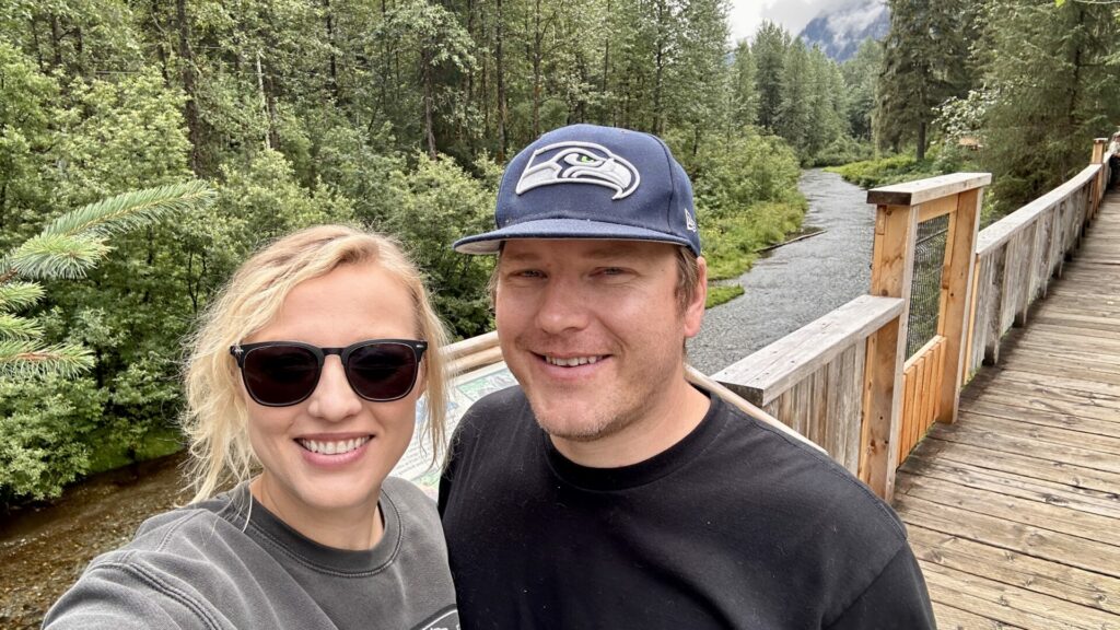 Rae and Jason of Getaway Couple standing on a boardwalk with a small river behind them. In the down of Hyder, Alaska, off of the Cassiar Highway. 