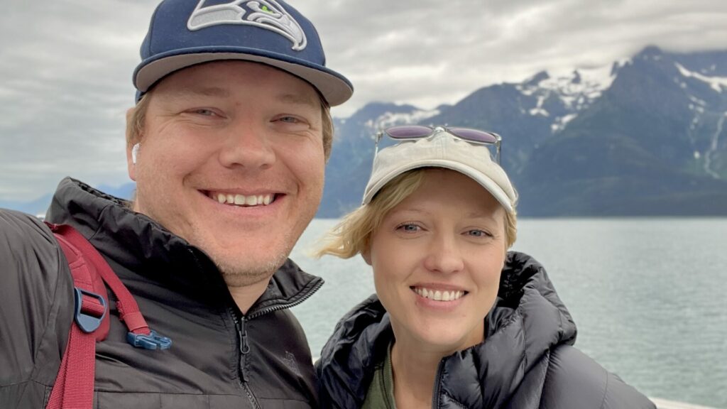 Rae and Jason smiling while on a ferry with water and snow covered mountains in the background. 