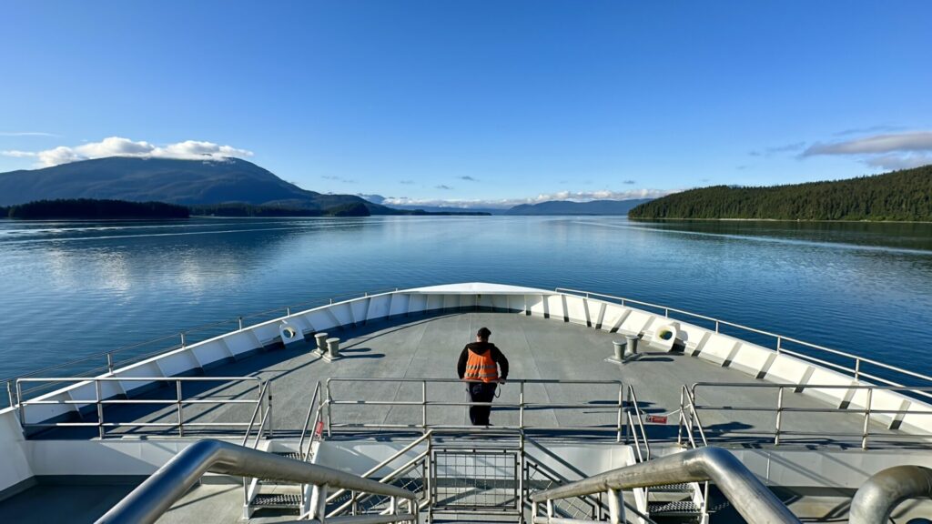 A man standing at the front of the ferry to Juneau, Alaska overlooking the blue ocean.