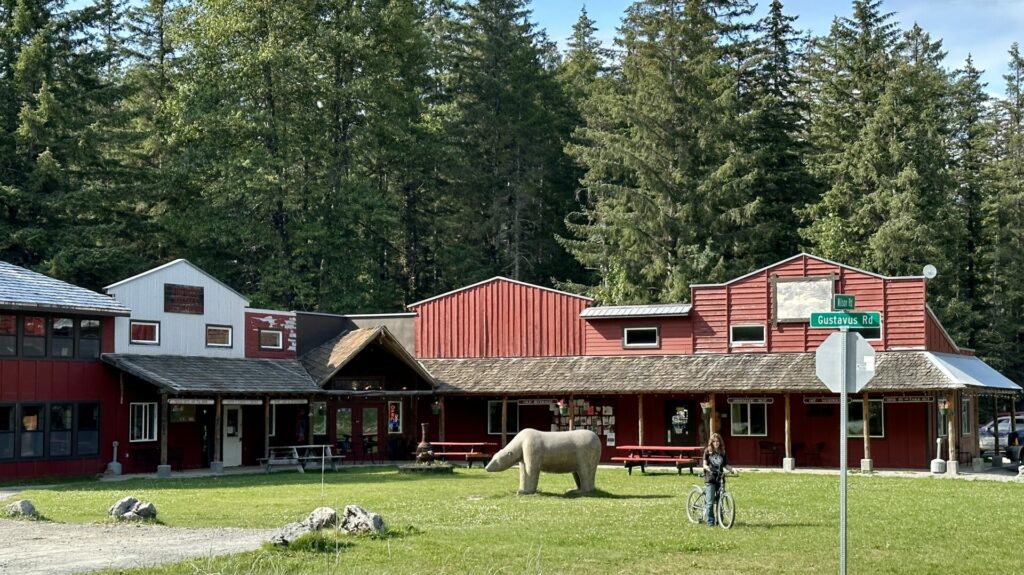 The small downtown area of Gustavus, AK showing the row of shops and a fake bear carved out of wood in the middle of the lawn.