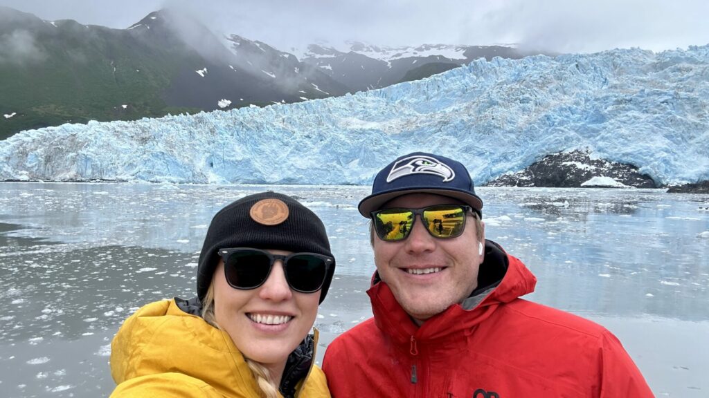 A couple smiling taking a selfie with one of the many glaciers in Alaska in the background. 