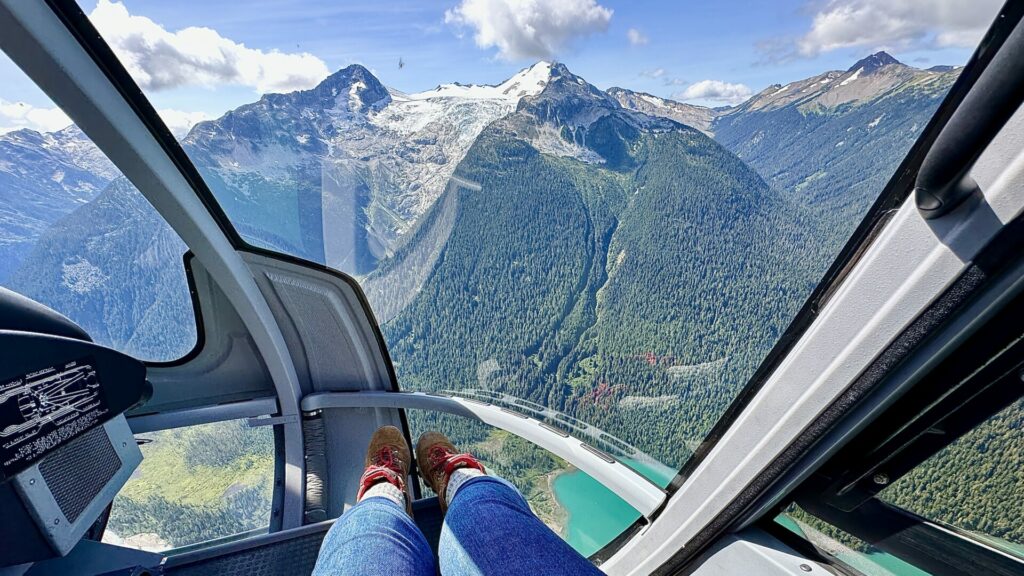 A woman's feet in a helicopter overlooking a lake and mountain range with a glacier on it. 
