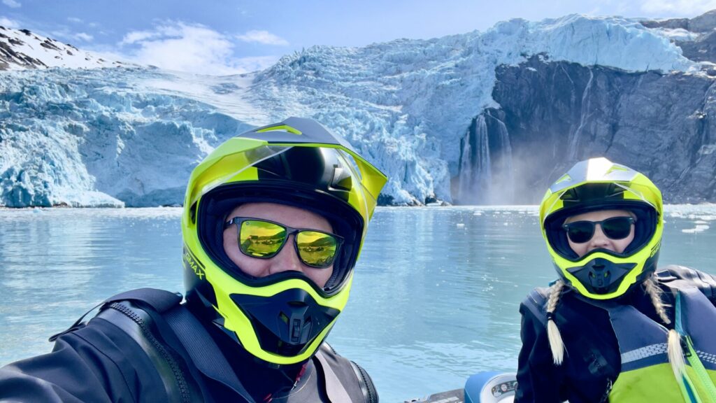 A couple on jet skis wearing yellow helmets taking a selfie in front of a glacier with a waterfall. 