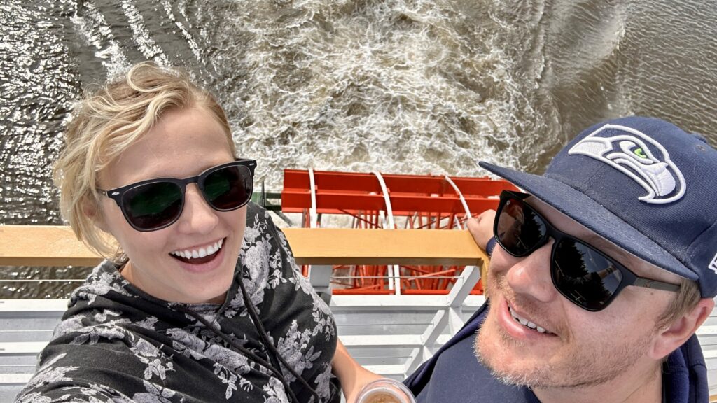 A man and woman standing at the rear of a paddle boat tour taking a selfie with it rolling in the water.