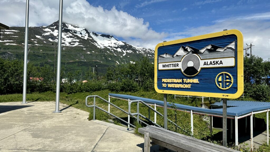 A blue sign that says Whittier Alaska with a mountain and blue skies. 