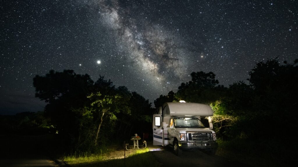 An RV parked in a campsite at night with the stars and milky way above. 