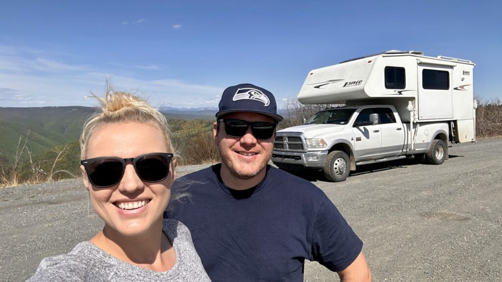 A young couple taking a selfie standing in front of their truck camper with a vast view behind it. 