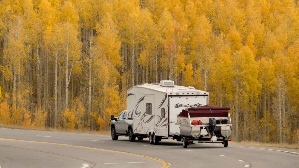 A truck towing an RV and a boat behind that, this is called triple towing. It is driving down the highway with bright yellow foliage in the background. 
