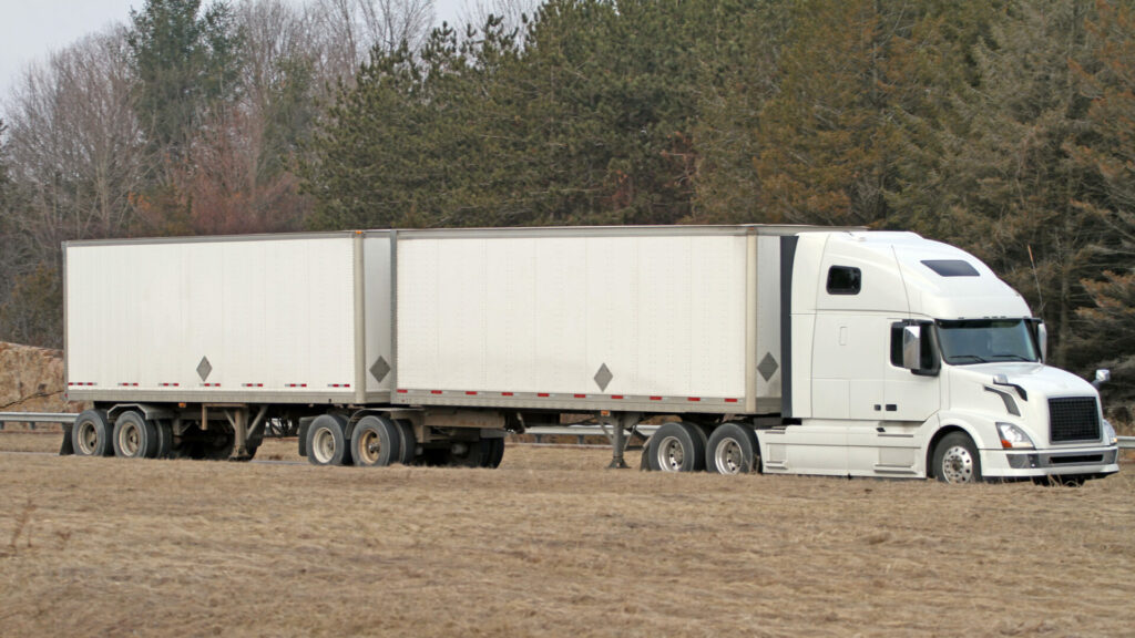 A white semi truck towing two trailers down the highway, this is called double towing or "hauling doubles" in the trucker community. 