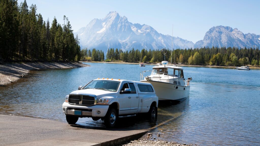 A white truck reversing on a boat ramp, loading his boat in the water. There is a beautiful mountain range in the background.