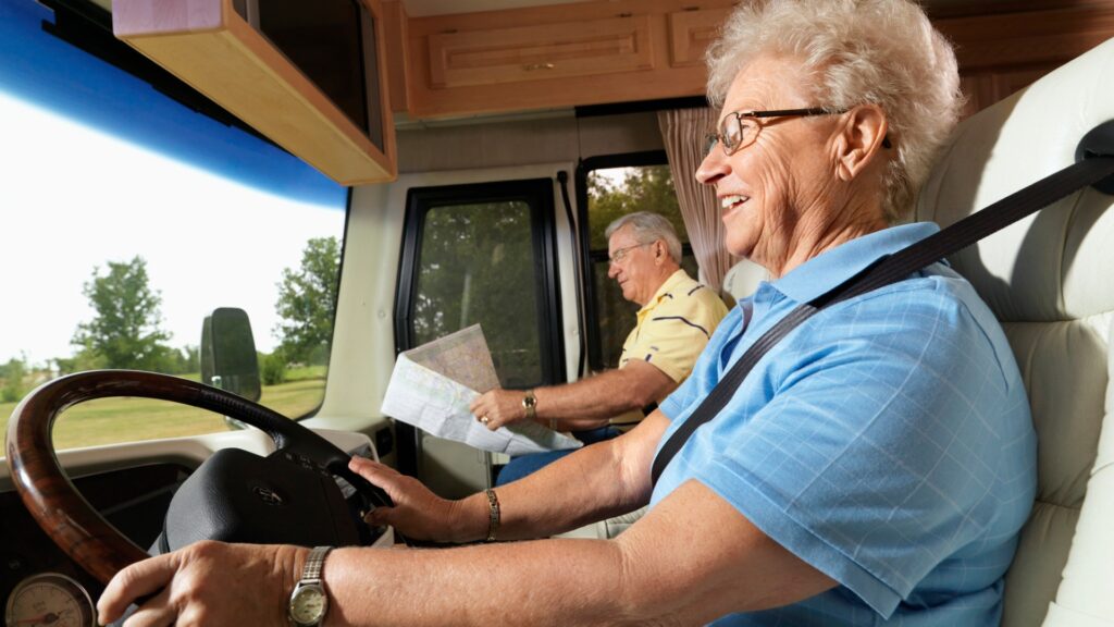 an elderly woman driving a motorhome while smiling and a male passenger looking at a map. 