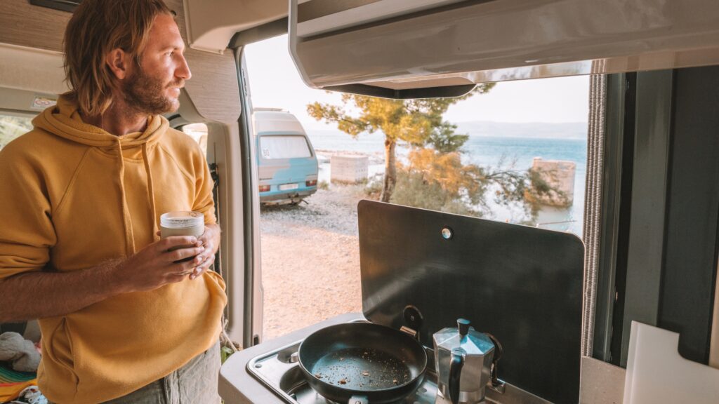 A man looking out the door of his RV while holding coffee and breakfast cooking in front of him. 