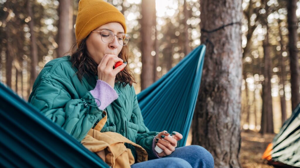 A woman sitting in a hammock using her inhaler due to camping with allergies.
