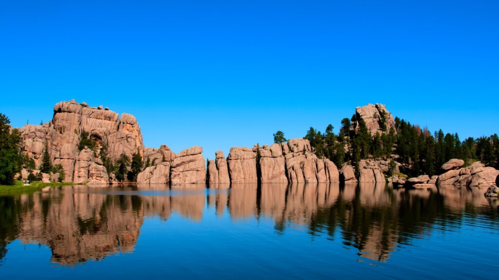 A reflective lake in Custer State Park with rocks in the background and a bright blue sky.