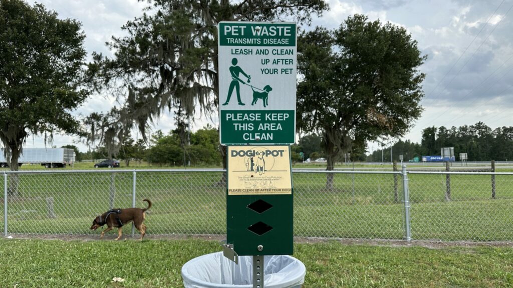 A sign that is asking people to clean up their dog waste in a campground with Carmen the doberman in the background