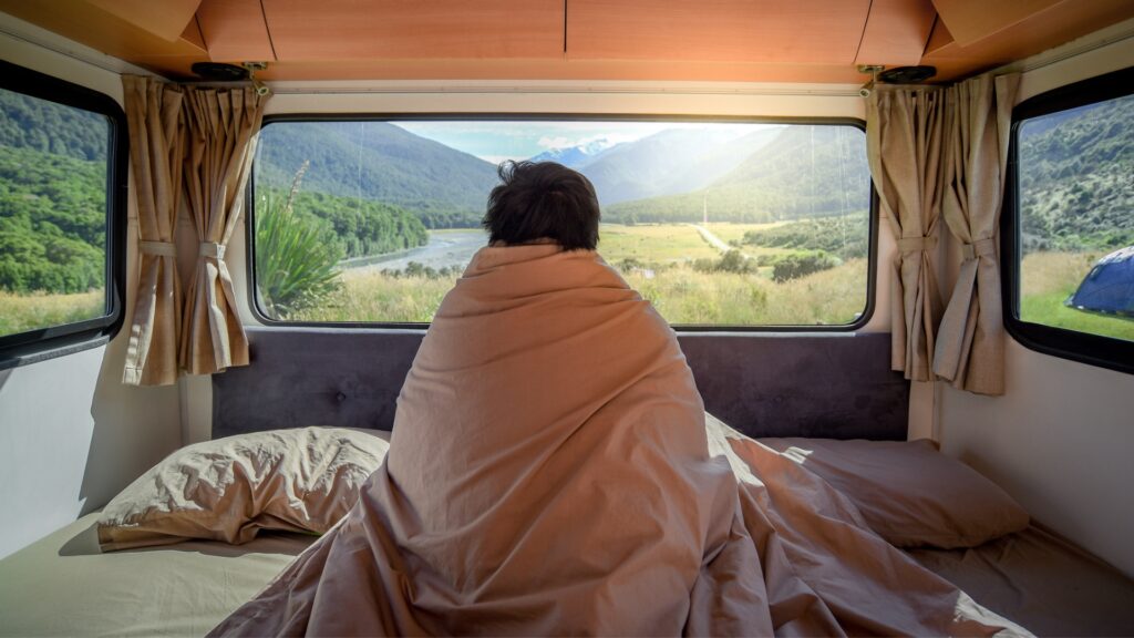 A person wrapped in blankets in an RV bed looking out the big windows at a pretty view.
