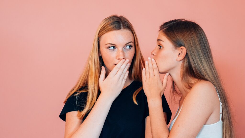 Two girls with their mouths covered gossiping in front of a pink wall.