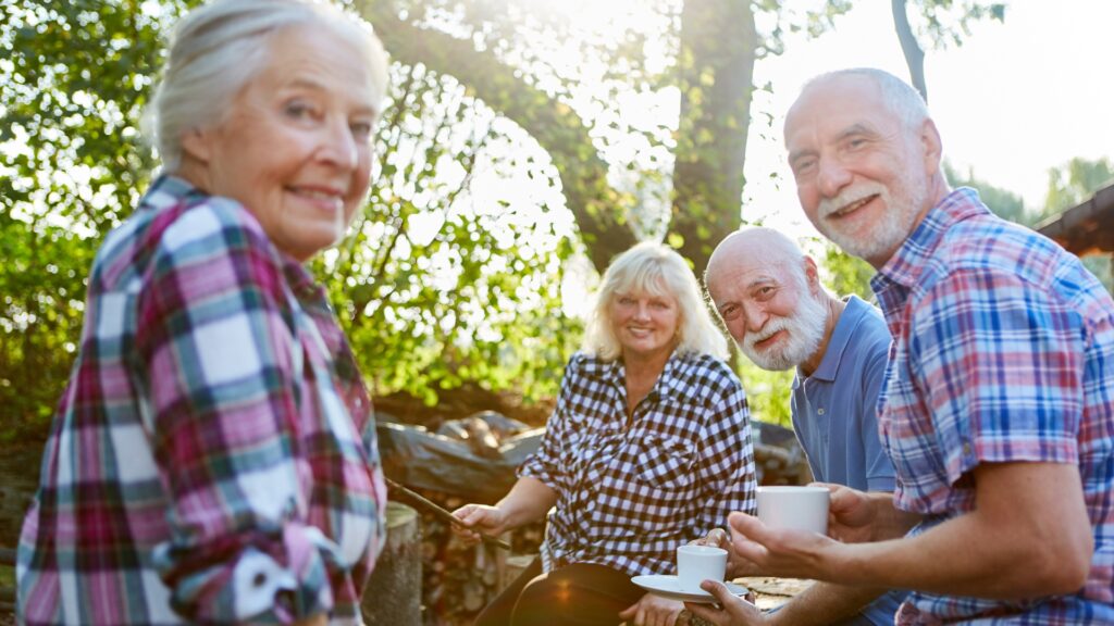 Four elderly people sitting in front of a campfire with coffee smiling at the camera. 
