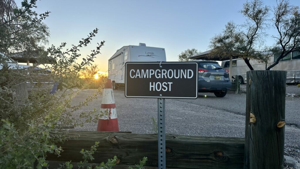 A sign that says campground host with an RV in the background. It's during sunset. 