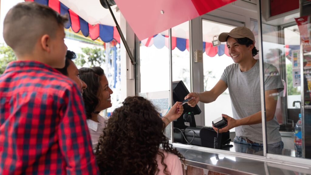 A young man working in a food truck at an amusement park handing back the credit card to the customer he is helping. 