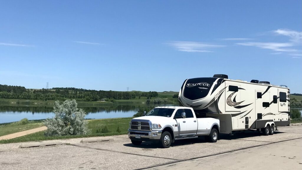 A fifth wheel being towed by a truck parked at a pullout with a pretty background of blue skies and water. 