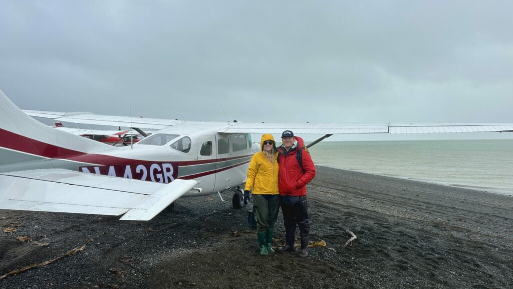 Jason and Rae, an adventure couple standing in front of a six seater plane that is on the beach in Alaska.