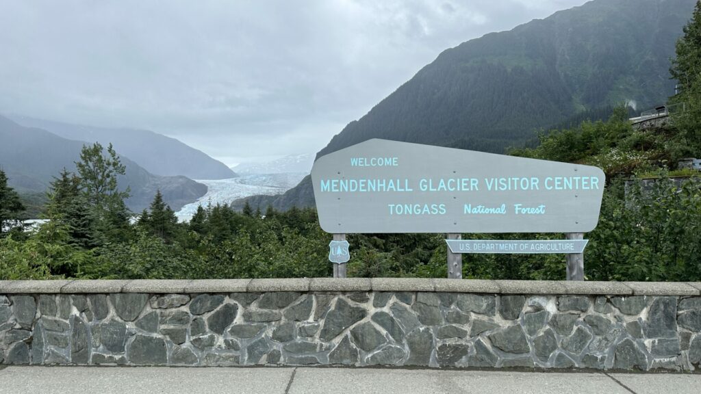 A sign that says "Mendenhall Glacier Visitor Center" with the Mendenhall Glacier in the background on a cloudy day.