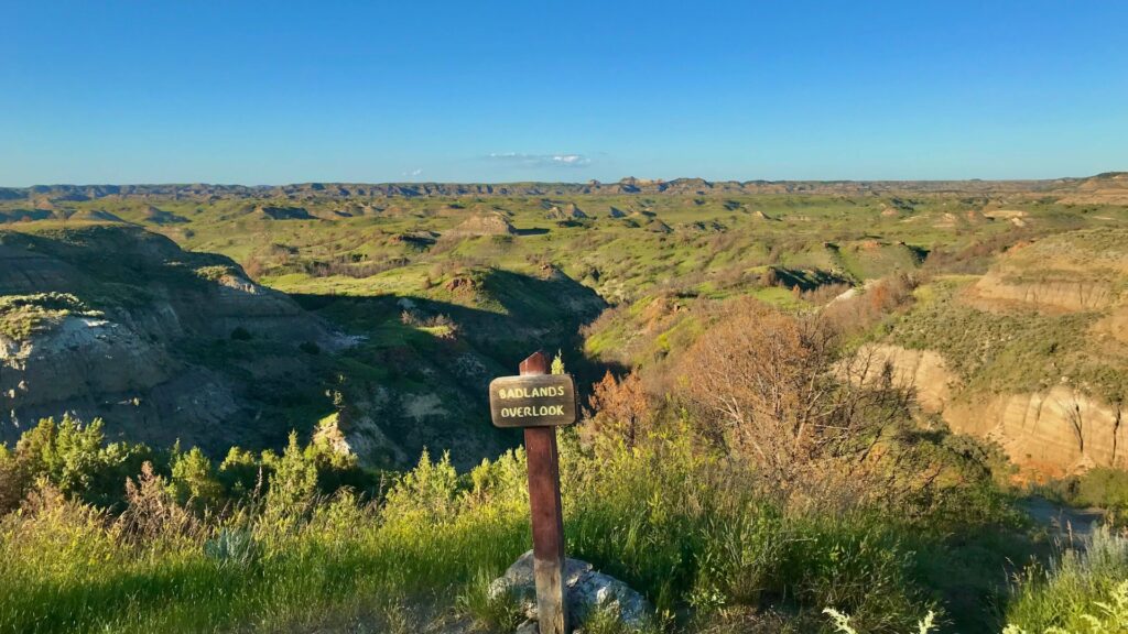 The Badlands Overlook sign with the badlands in the background covered in green due to the spring season. 