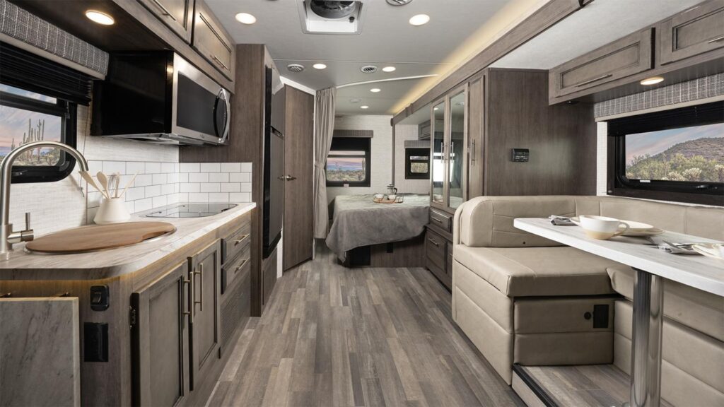 Interior shot of a Jayco Melbourne Class C RV. Brown cabinets and floor with a u-shaped dinette and bed in the back of the RV. 