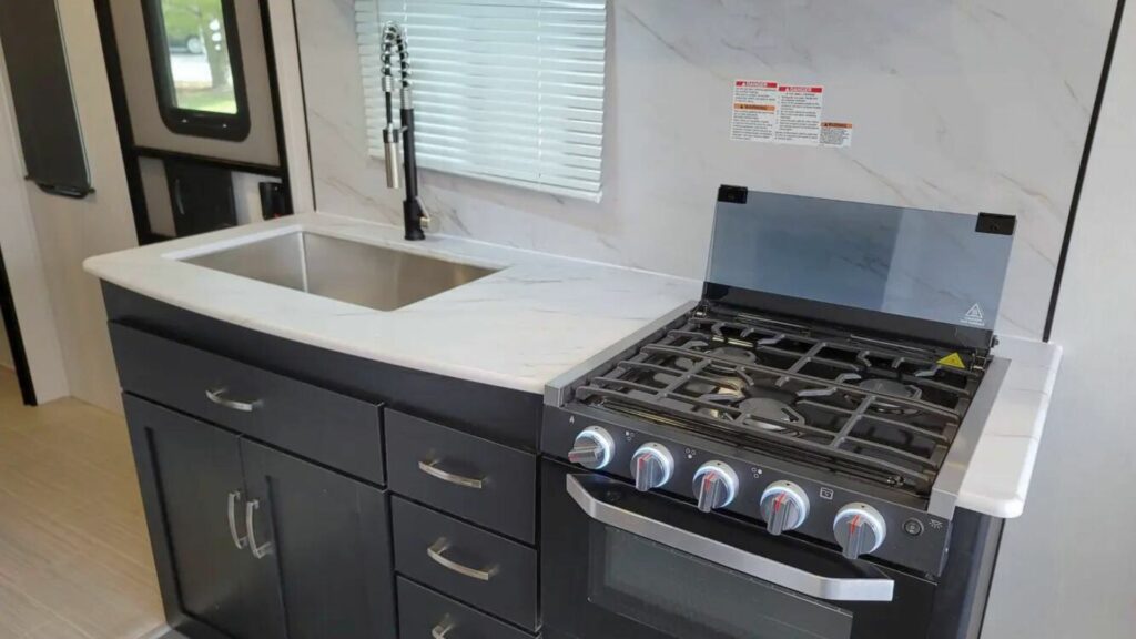 The kitchen area of a Work and Play toy hauler showing black cupboards, a white counter, black stove, and stainless steel sink.