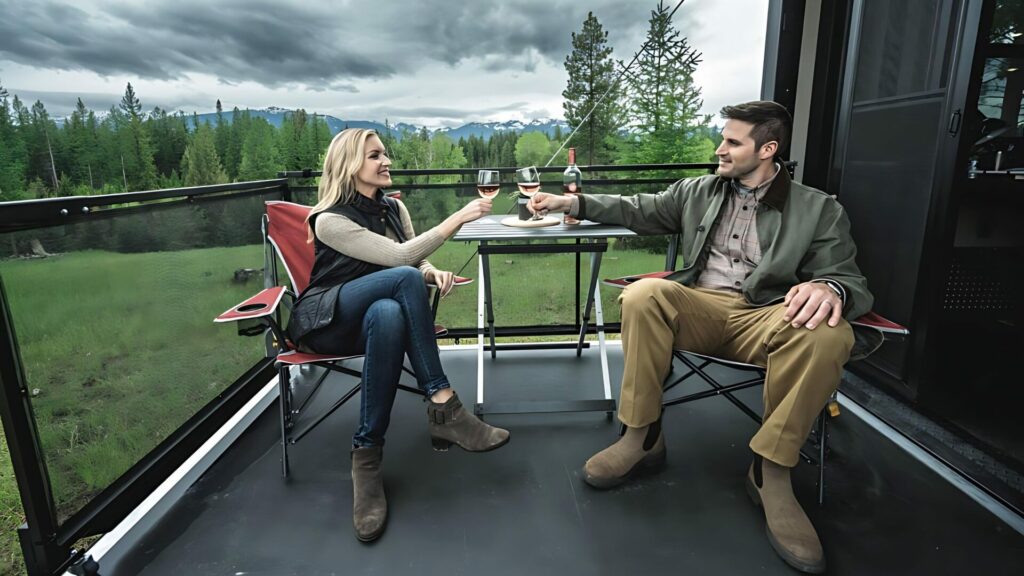 Man and woman sitting in camping chairs, drinking wine as they sit on a D RV Patio with a mountain range in the background. 