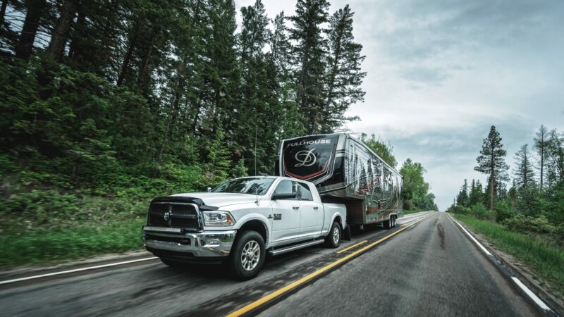 White truck pulling DRV Toy Hauler on the road with forest in the background.