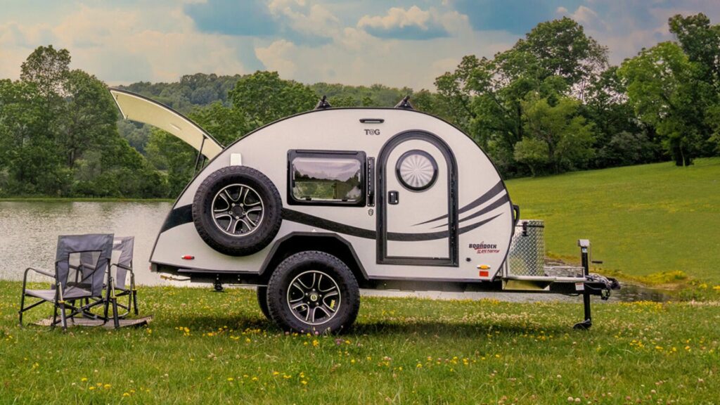 Tag trailer camper by nuCamp in front of a lake with camping chairs.