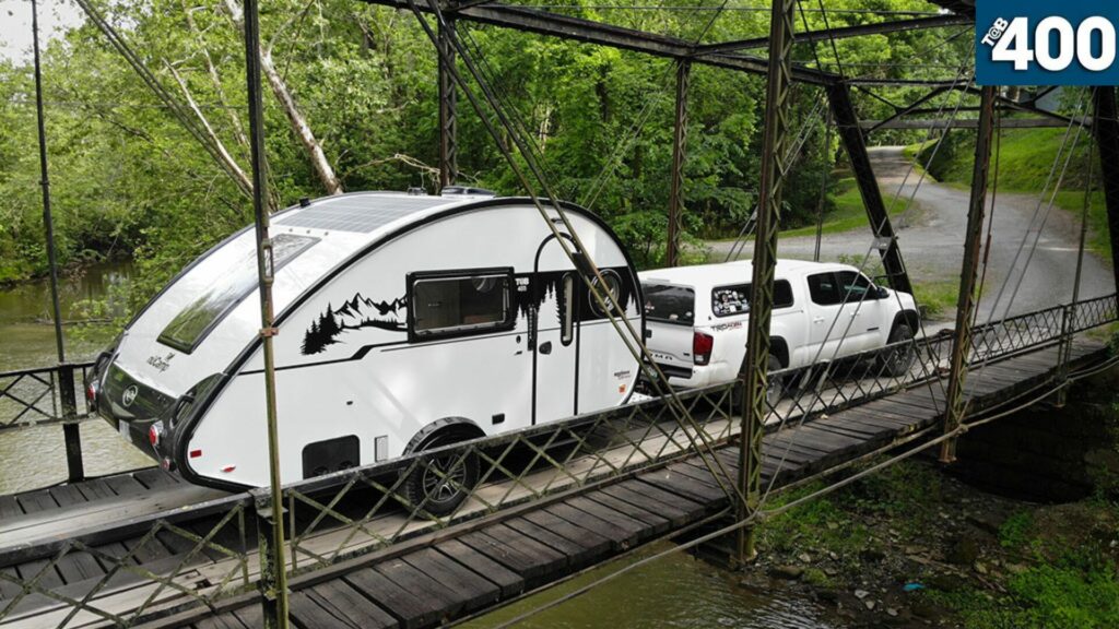 The nuCamp Tab 400 teardrop trailer being towed by a white pickup truck over a heavily wooded bridge. 