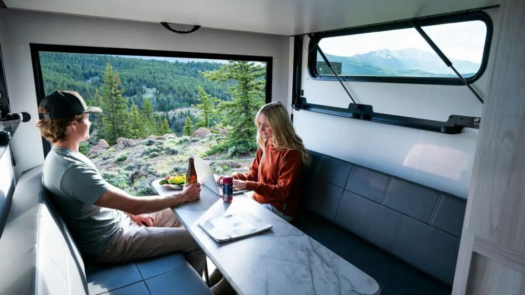 A couple sitting at the dinette inside their Winnebago Hike 100 travel trailer with a heavily wooded forest view. 