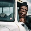 Person dressed up as bigfoot driving a white pickup truck as they lean out the window.