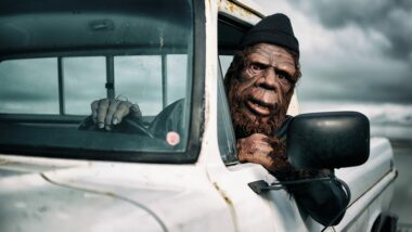 Person dressed up as bigfoot driving a white pickup truck as they lean out the window.