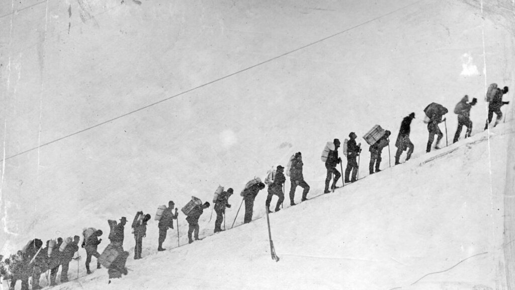 People with gear on their backs stand in a single file line headed uphill with a snowy backdrop. Stampeders, loaded with gear, wait in line starting up Chilkoot Pass