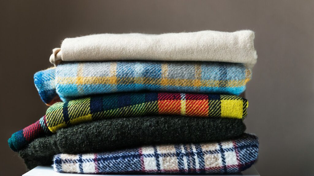 A stack of five cashmere wool blankets in assorted colors and varying in plaid and solid patterns. They sit folded and stacked atop a table with a warm grey background.