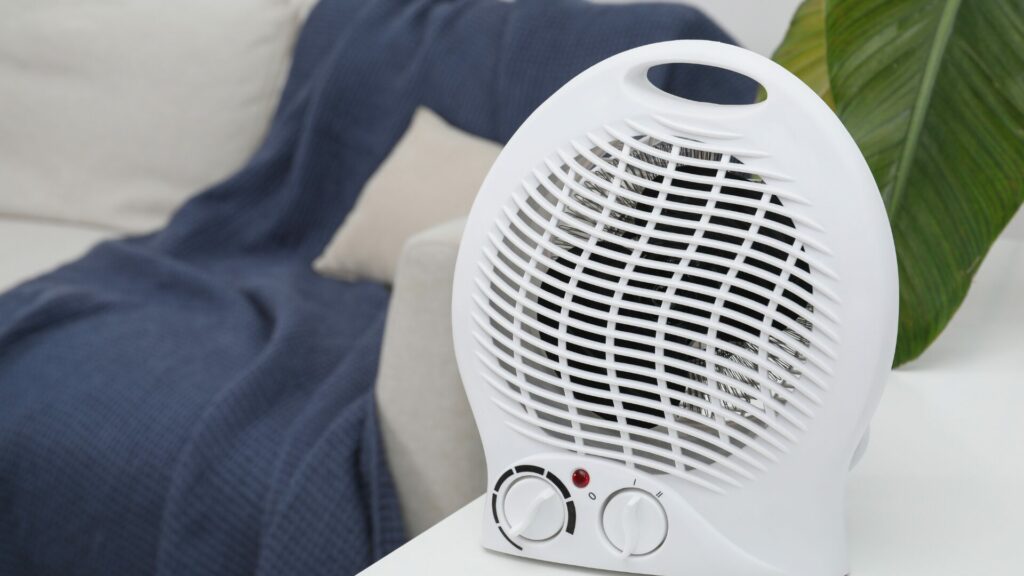 A small white portable heater on a table next to a couch with a throw blanket and a tropical plant in the background.