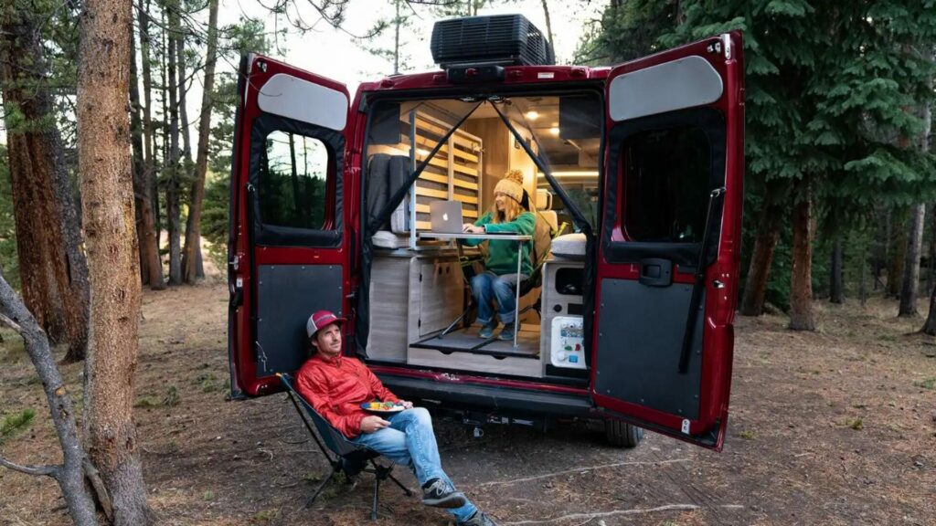 The Winnebago Solis set up with a woman working inside and a mansitting in a camping chair outside. 