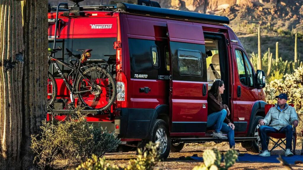 The Winnebago Travato 59G in a desert with a woman sitting on the floor of the doorway and a man sitting in a camping chair. 