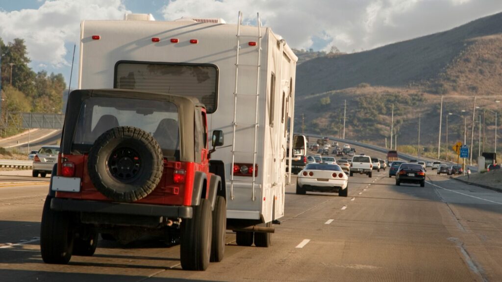 A red Jeep is being flat-towed by a class C motorhome on the highway. 