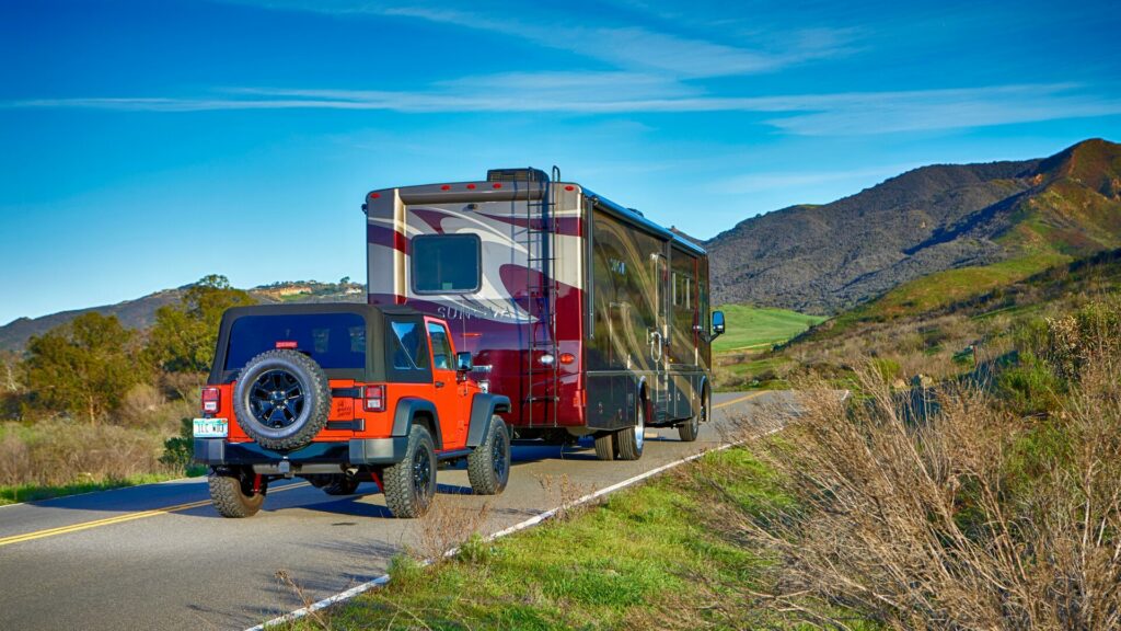 A Class A motorhome flat towing a red jeep through some hills on a paved road. 