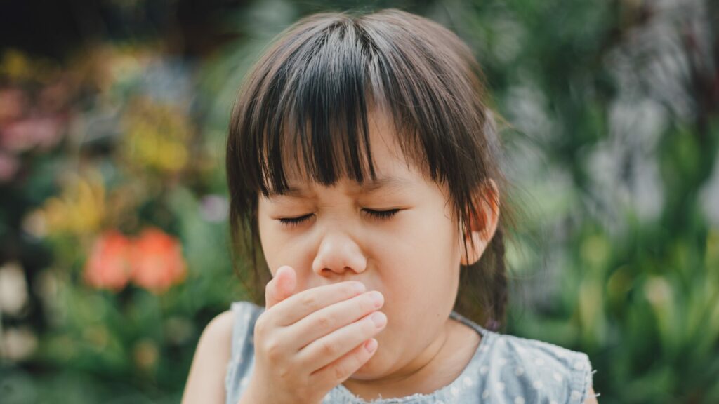 A small child coughing as they hold their hand over their mouth with their eyes closed while standing outside. 