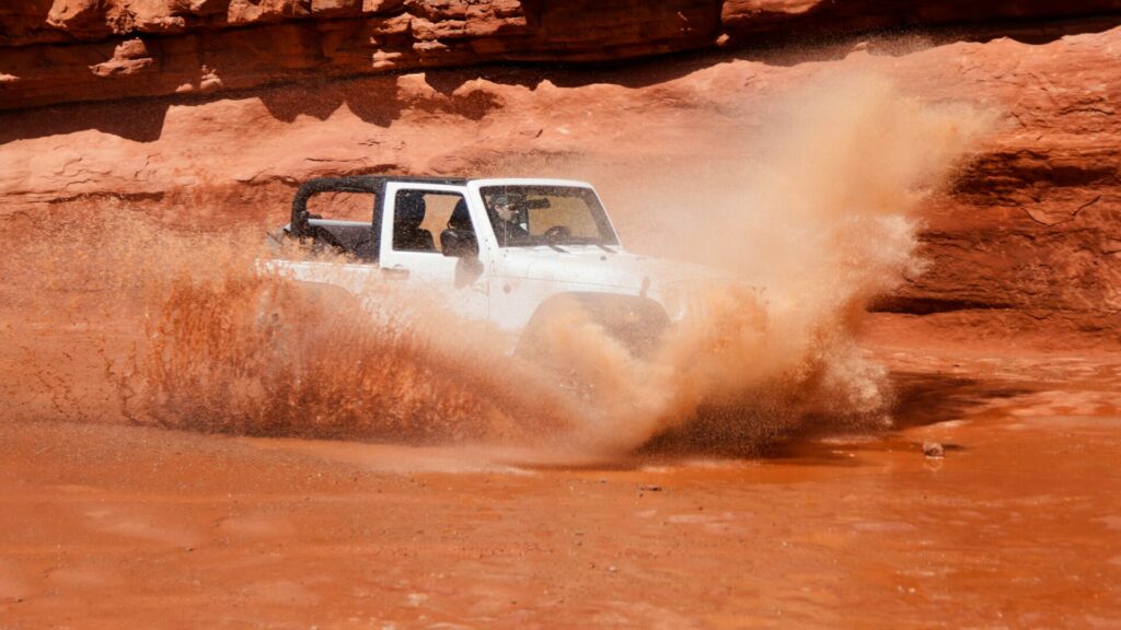 A white vehicle driving through a red-orange mud pit, causing the mud to splash upwards. 