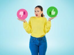 Woman in yellow sweater gesturing "I don't know" with a pink thumbs down in one hand and a green thumbs up in the other.