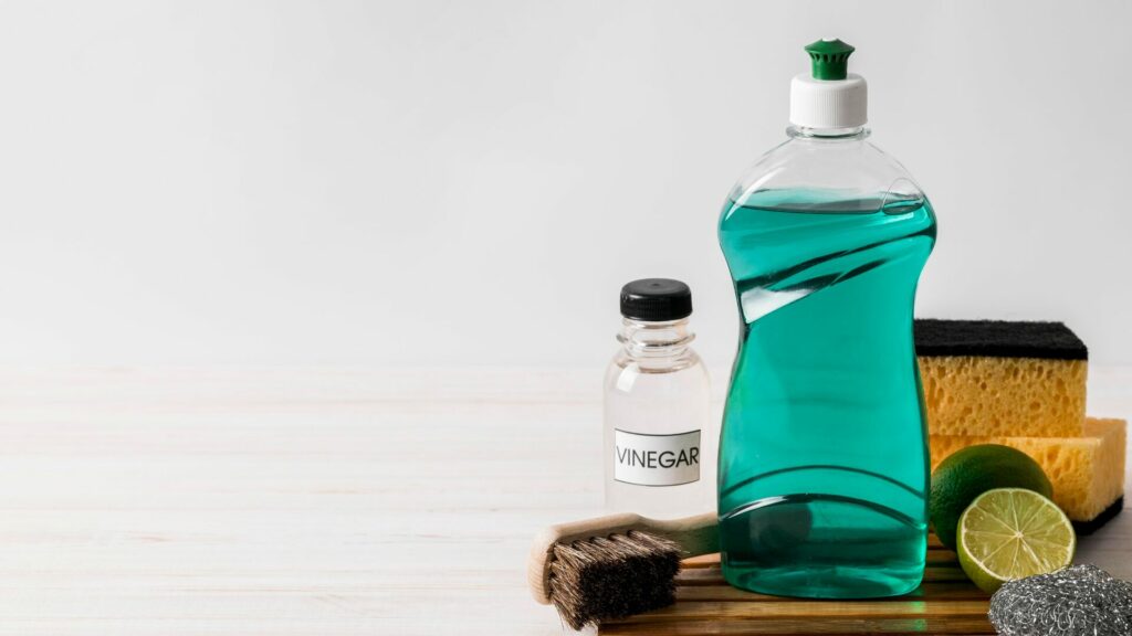 A wood plank with a brush, steel wool, a lime, dish soap, and a bottle of vinegar sitting atop it with yellow sponges in the background. 